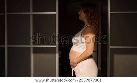 A pregnant woman in shorts and a crop top stands in profile between folding doors.
