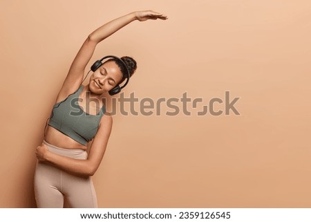 Horizontal studio shot of slim sporty woman leans aside stretches arms listens music via headphones dressed in cropped top and leggings isolated over brown background copy space for your advertisement