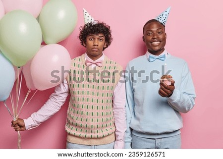 Horizontal shot of curly haired Hindu man and black man dressed in festive clothing holds cupcake with candle holds bunch of colorful balloons celebrates special occasion isolated over pink background Royalty-Free Stock Photo #2359126571