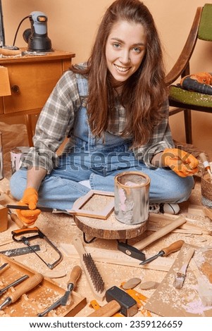 Shot of young pretty happy smiling European woman wearing denim jumpsuit and shirt sitting on floor of handicraft workshop painting picture frame looking straight at camera. Handmade concept