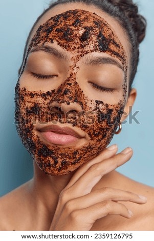 Beauty and healthy skin concept. Calm young dark haired woman touches face gently applies facial coffee scrub for cleaning skin keeps eyes closed stands shirtless isolated over blue background