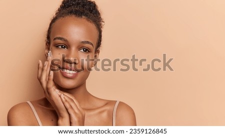 Pleasant looking woman applies day cream to keep skin collagen and elastin rate hydrate and nourish it has tender expression smiles gently isolated on brown background with blank space for your promo Royalty-Free Stock Photo #2359126845