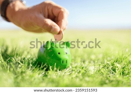 Hand inserting coin and saving money in green piggy bank with grass and blue sky background, savings, accounting, banking and business account or sustainable and environmentally friendly finance Royalty-Free Stock Photo #2359151593