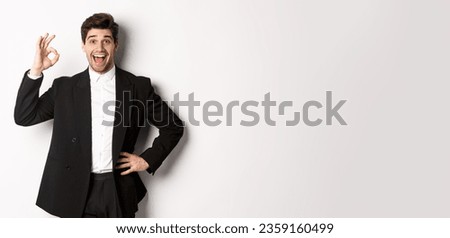 Concept of new year party, celebration and lifestyle. Portrait of pleased handsome guy in black suit, praise something good, showing okay sign in approval, standing over white background.