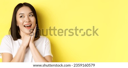 Cute romantic girl in white t-shirt imaging valentines day, looking left with admiration and joy, smiling with hands pressed to cheek, standing over yellow background.