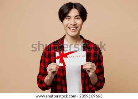 Young satisfeid happy man of Asian ethnicity wear red shirt casual clothes hold gift certificate coupon voucher card for store isolated on plain pastel light beige background studio. Lifestyle concept