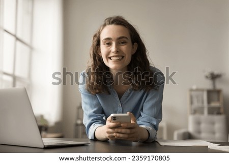 Happy young gen Z freelancer woman using Internet technology for job business communication, working at home, sitting at laptop, holding smartphone, smiling, laughing, looking at camera for portrait Royalty-Free Stock Photo #2359170805