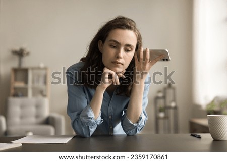 Serious freelancer employee girl holding mobile phone at head, listening to sound, voice message, audio file, leaning hands on home workplace table, thinking, touching chin Royalty-Free Stock Photo #2359170861