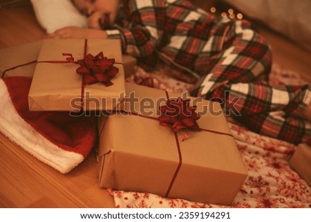 In the foreground close-up there is a craft gift boxes with a red bow in the background there is a sleeping child in plaid pajamas