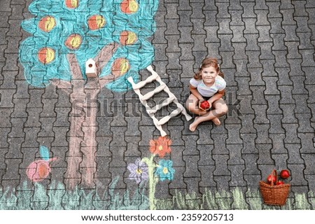 Cute little girl picking apples from apple tree painted with colorful chalks on asphalt. Cute preschool child with having fun with chalk picture. Creative leisure for children, drawing and painting.