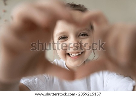 Positive sweet little preschool girl looking at camera through hand heart frame with toothy smile. Cheerful pretty little kid joining fingers, showing gesture sign of happiness, love, happy childhood