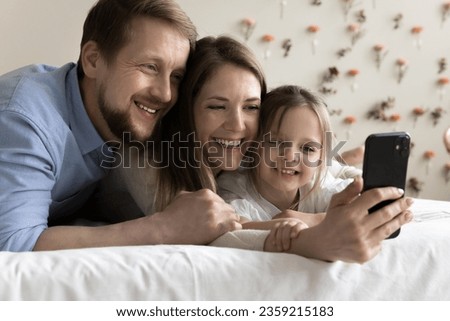 Happy young parents hugging little toddler kid, talking selfie on cellphone, posing for self picture together, speaking on online family video call, enjoying domestic Internet communication