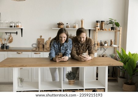 Positive digital addicted couple standing close in kitchen, using gadgets separately, avoiding offline talk, communication, reading social media content, chatting mobile phone