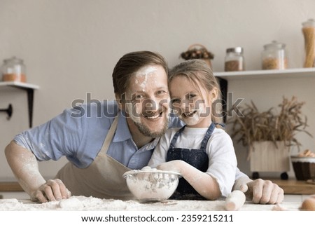 Cheerful funny little daughter and dad posing in home kitchen with floury faces, looking at camera, laughing, standing at table with mess of flour powder, enjoying baking, family culinary hobby