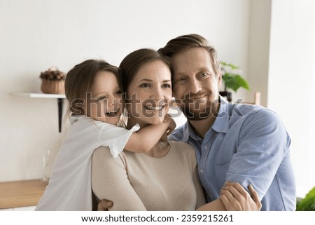 Cheerful little daughter girl and young handsome dad hugging pretty joyful mom with love, tenderness, celebrating mothers day, looking away, posing for family shooting together at home