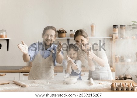 Joyful excited parents and little kid having fun at bakers kitchen table, making mess, clapping floury hands over counter, looking at camera through cloud of flour, baking, laughing Royalty-Free Stock Photo #2359215287