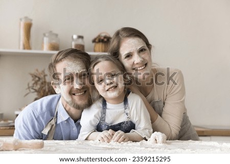 Happy young mom, dad and toddler kid in aprons with funny floury facial masks cooking dessert in home kitchen, having fun, posing at table with heap of flour, looking at camera, laughing