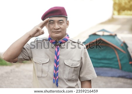 Handsome Asian man wear boy scout uniform red cap blue and pink striped scarf, make hand sign symbol at outdoor campingsite.Concept, educational career with uniform in school, Thailand. Go camping    