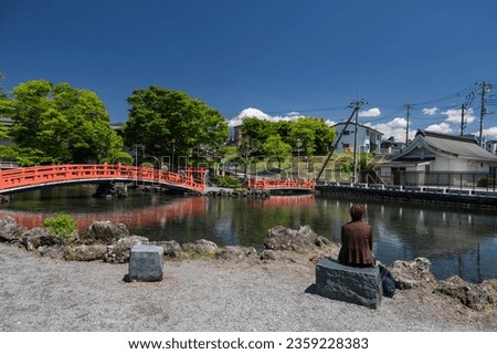 Old Japanese woman sit and enjoy view of red bridge over pond and Fujinomiya city and mount Fuji against blue sky, Shizuoka, Japan. Famous travel destination. Royalty-Free Stock Photo #2359228383