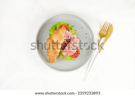 burger croissant with sausage, green, tomato, knife and spork on grey plate on a light background, top view