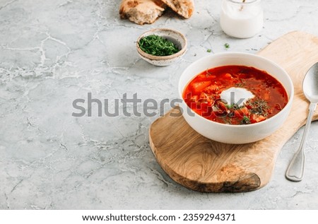 Delicious traditional Ukrainian and Russian borscht served in a white bowl on a grey marble background. Accompanied by a sauceboat filled with dill, bread, and a jar of sour cream Royalty-Free Stock Photo #2359294371