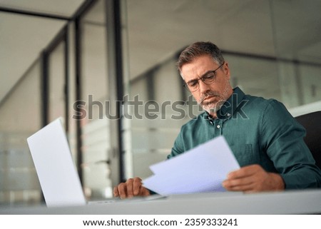 Busy middle aged executive, mature ceo, male hr account manager holding documents using laptop computer in office, checking financial bill, data in report, doing accounting paper file plan overview. Royalty-Free Stock Photo #2359333241