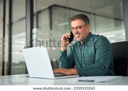 Busy smiling older middle aged business man professional expert or entrepreneur making phone call speaking with client communicating on cellphone using laptop computer sitting at desk in office.