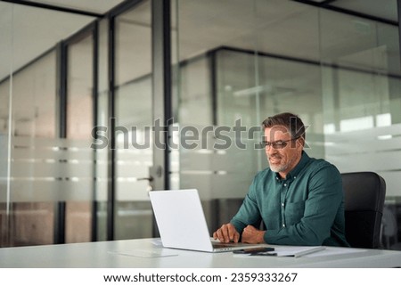 Smiling busy older professional business man working on laptop sitting at desk. Older mature Indian businessman, happy male executive manager typing on computer using pc technology in office.