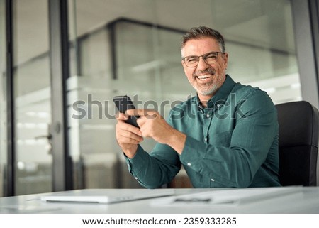 Cheerful older professional businessman, happy middle aged business man entrepreneur laughing looking at camera holding smartphone using cell phone mobile technology sitting at work desk in office. Royalty-Free Stock Photo #2359333285