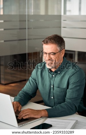 Smiling busy mid aged professional business man working on laptop at office desk. Older mature Indian happy male entrepreneur worker typing on computer, executive manager using pc in office. Vertical