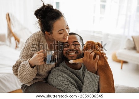 Positive interracial couple in cozy sweaters holding coffee while playing with chihuahua dog at home Royalty-Free Stock Photo #2359336545