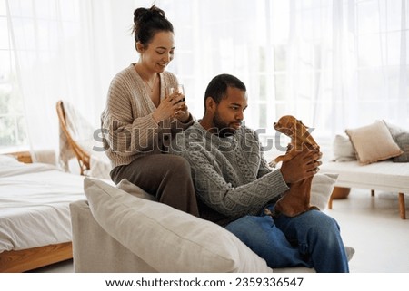 Joyful young multiethnic couple in warm clothes spending time with dog and coffee at home