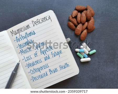 Symptoms and signs of magnesium deficiency written on the notebook, with almond and pills in background. Low magnesium level may cause weakness, fatigue, osteoporosis, muscle spasms, arrhythmia, etc. Royalty-Free Stock Photo #2359339717