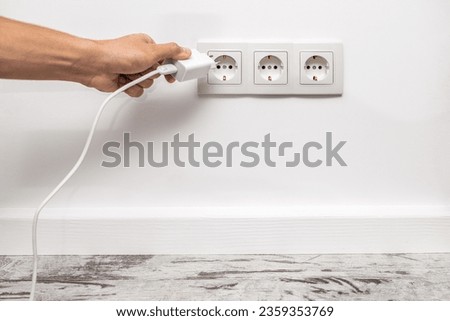 The human hand unplugging a phone adapter from a triple white electrical outlet, situated on a white wall. Royalty-Free Stock Photo #2359353769