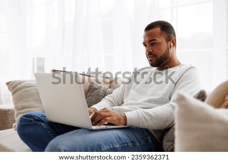 African american freelancer in wireless earphone working while using laptop on couch at home Royalty-Free Stock Photo #2359362371