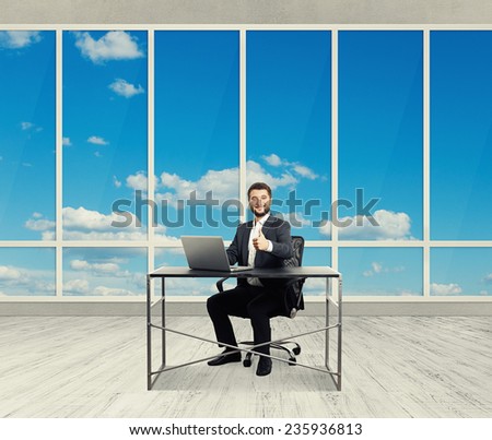 smiley successful businessman sitting at the table with laptop, showing thumbs up and looking at camera in the light office with big windows