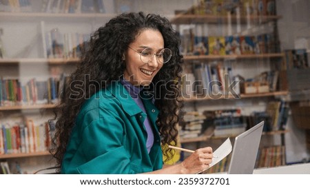 Cheerful young Oriental woman tutor freelancer student writing at copybook homework test, e-learning, working remotely, watching webinar on laptop. Education learning concept on bookshelves background