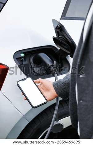 Businessman traveling by electric car having stop at chraging station standing holding smartphone looking at screen copy space for text or product close-up while having vehicle fully charged Royalty-Free Stock Photo #2359469695