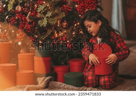 Happy japan little girl dreamer waiting miracle from Santa posing on floor near noel tree. Asian kid in red plaid outfit celebrating new year with gifts boxes at bokeh xmas lights eve 25 december