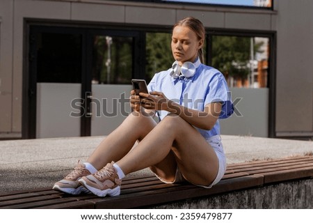 Stylish woman university student with headphones using smartphone app sitting on bench outdoors online learning, remote studying virtual class, watching webinar distance course or listening podcast.