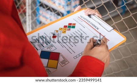 A worker is checking on the hazardous chemical material information form with background of chemical storage area at the factory place. Industrial safety working action. Selective focus.	
