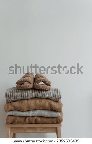Warm female home wearing. Stack of neutral colour sweaters, pullovers and fluffy slippers on wooden stool over white wall. Autumn, fall styled fashion composition Royalty-Free Stock Photo #2359505405