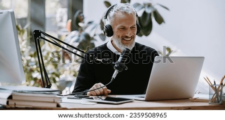 Cheerful mature podcaster in headphones using microphone while broadcasting from studio