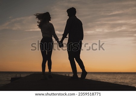 Silhouette of happy couple holding hands while standing at the sea dock. Two romantic people enjoying the sunset at beach.