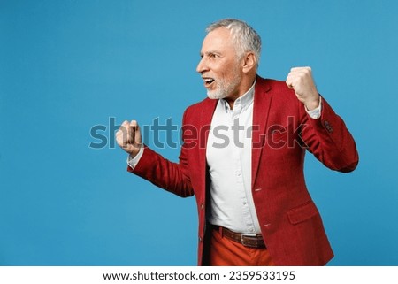 Excited elderly gray-haired mustache bearded business man wearing red jacket suit standing doing winner gesture clenching fists looking aside isolated on blue color wall background studio portrait Royalty-Free Stock Photo #2359533195