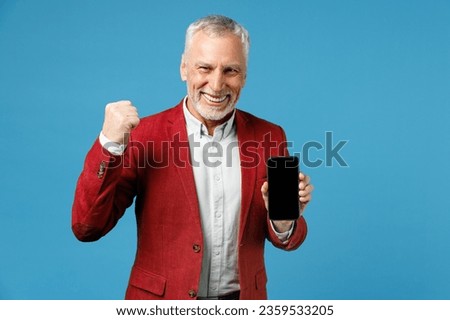 Happy joyful elderly gray-haired mustache bearded business man in red jacket suit hold mobile cell phone with blank empty screen doing winner gesture isolated on blue color background studio portrait Royalty-Free Stock Photo #2359533205