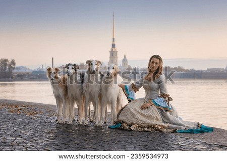 Elegant Russian Lady in Historic Dress with Borzoi Dogs near Neva River and Peter-Paul Fortress, St. Petersburg. Serene Morning, Clear Autumn Sky, Riverside Beauty Royalty-Free Stock Photo #2359534973