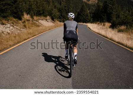 Close-up back view of a woman cyclist training on a mountain road. Cycling in the woods.Professional female cyclist is riding on a forest asphalt road.Fit athlete wearing sportswear and helmet.Romania Royalty-Free Stock Photo #2359554021