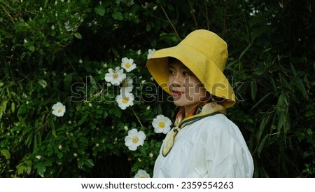 Portrait of pretty Asian woman in yellow hat and white shirt standing by white flower tree in spring field outdoor. 