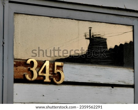 345, golden numbers. Home address. Reflection of the chimney in the glass. Wooden , white paint background. Sunrise. We learn to count, concept. Metal gray frame door.